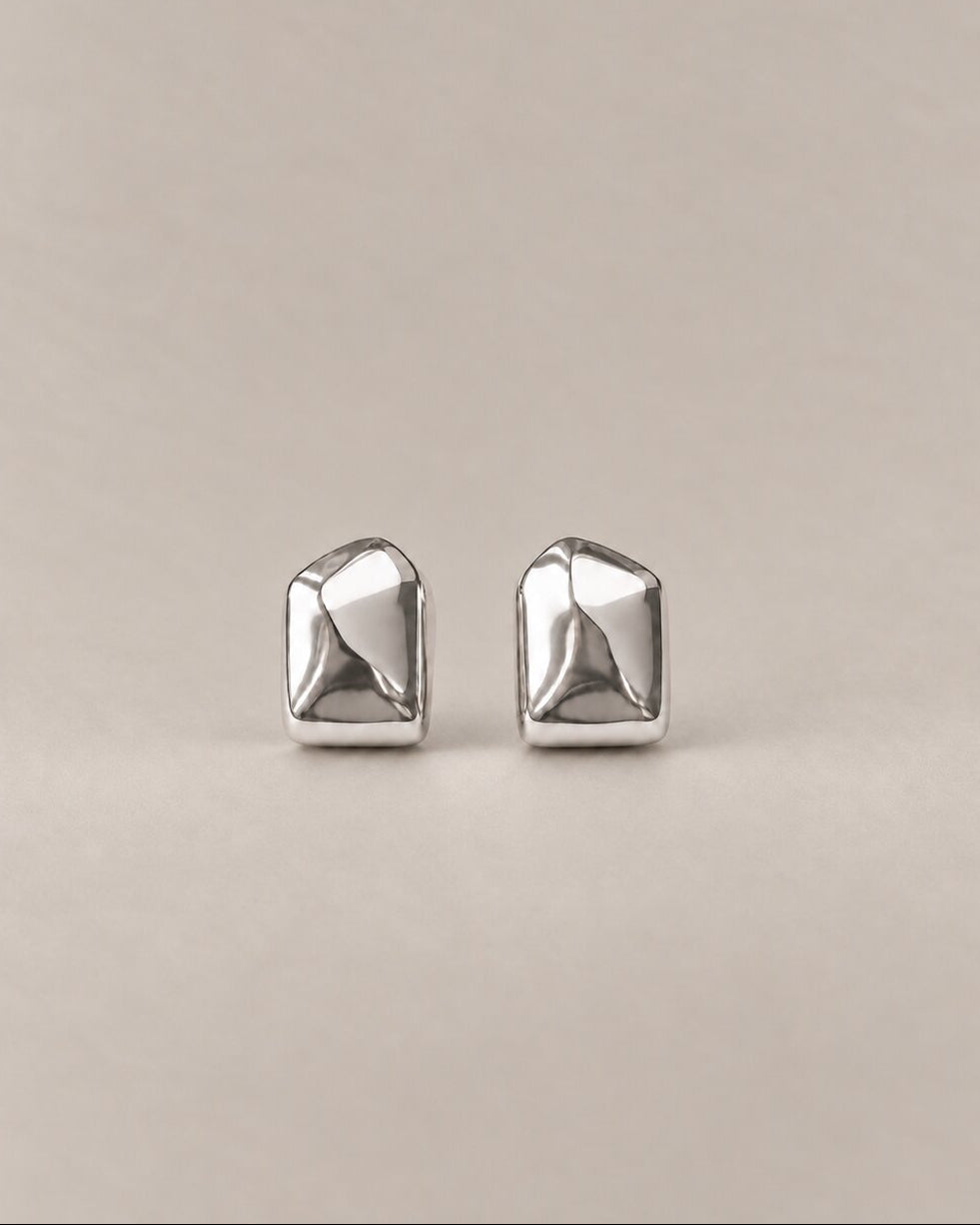 A pair of silver geometric stud earrings with a faceted, multi-sided design, displayed on a neutral beige background.