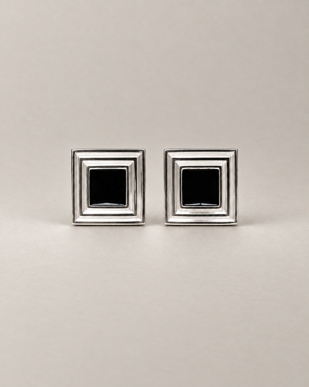 A pair of square silver-toned cufflinks with a black square inset in the center of each, displayed on a neutral light gray background.