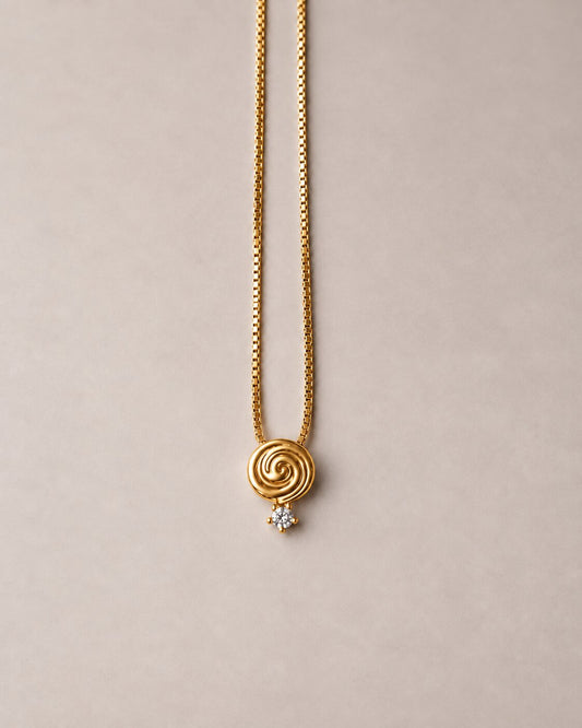 A gold chain necklace featuring a spiral-shaped pendant with a small clear gemstone accent, displayed against a solid beige background.