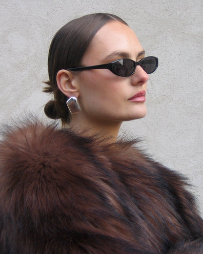 A woman in profile wearing black cat-eye sunglasses, a brown fur collar, and a silver geometric earring, set against a neutral gray textured wall.