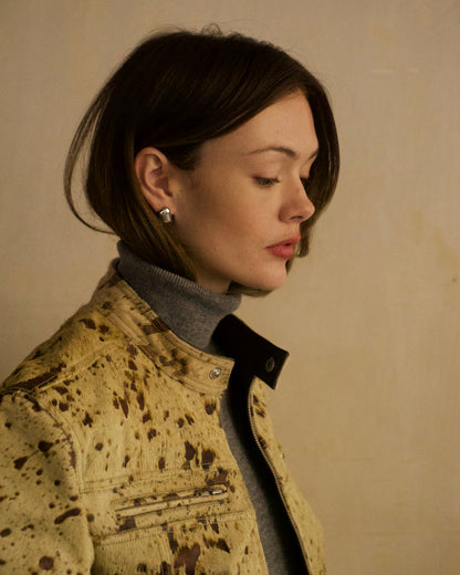 A woman in profile wearing a beige splatter-print jacket over a gray turtleneck, set against a plain beige wall.