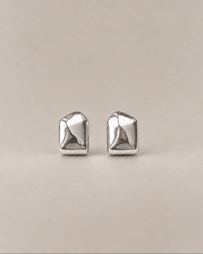 A pair of silver geometric stud earrings with a faceted, multi-sided design, displayed on a neutral beige background.