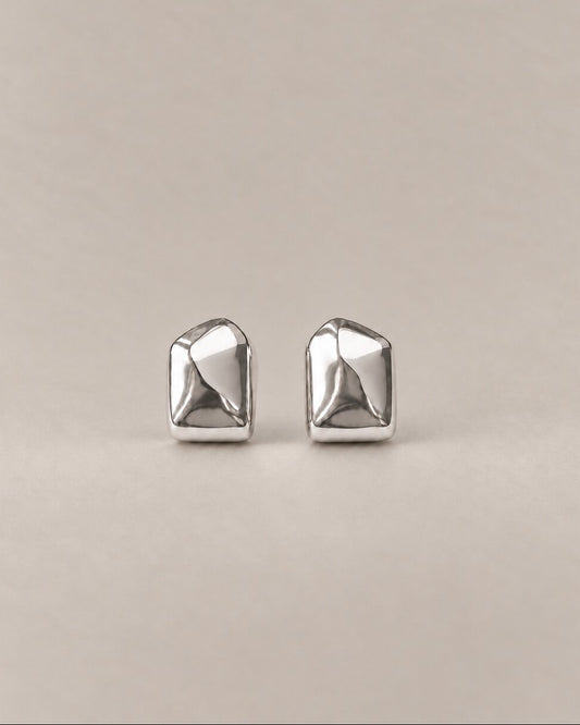 A pair of silver geometric stud earrings with a faceted, multi-sided design, displayed on a neutral beige background.