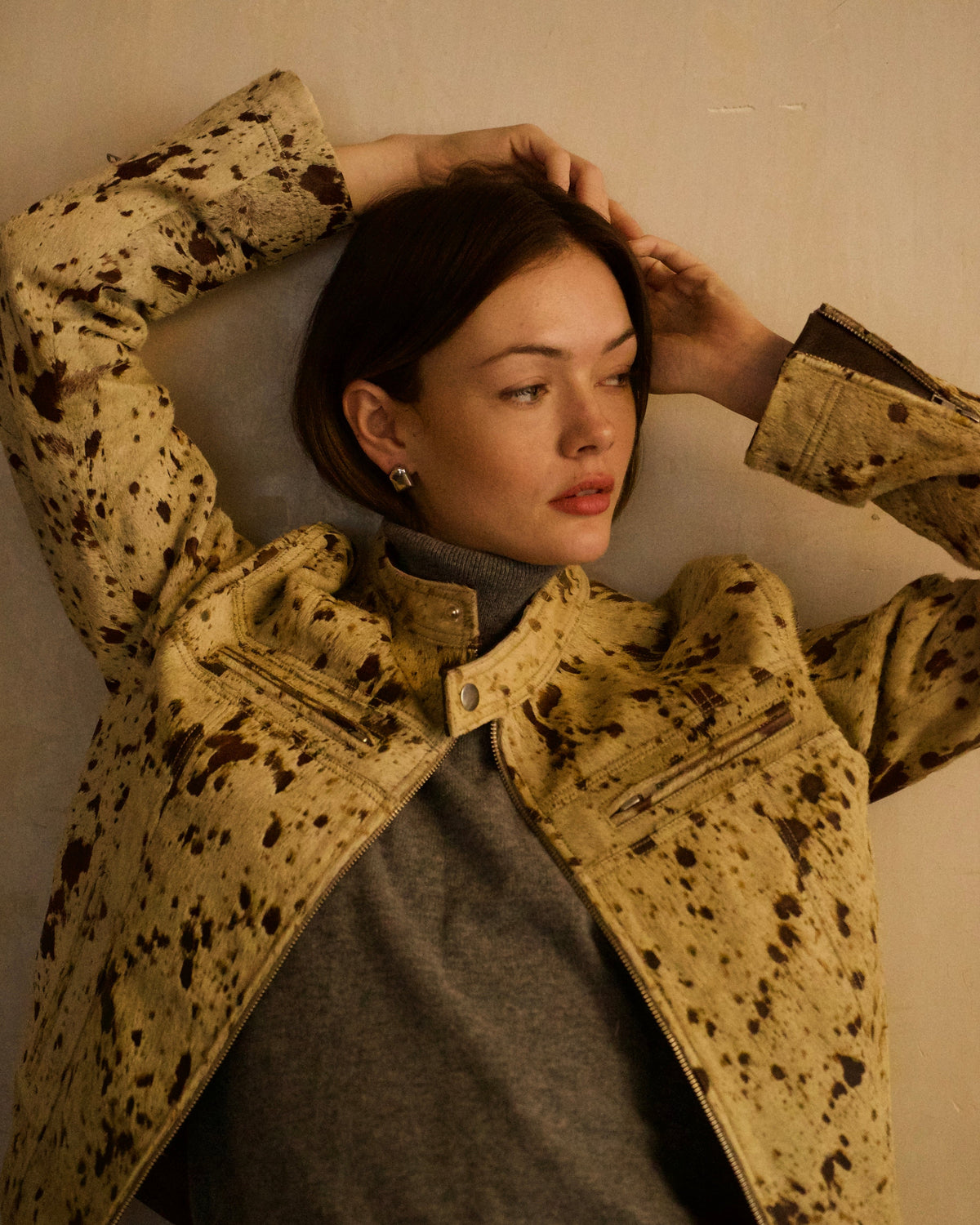 A woman wearing a beige and brown splatter-patterned jacket over a gray turtleneck sweater, posing against a textured beige wall.