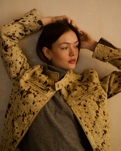 A woman wearing a beige and brown splatter-patterned jacket over a gray turtleneck sweater, posing against a textured beige wall.