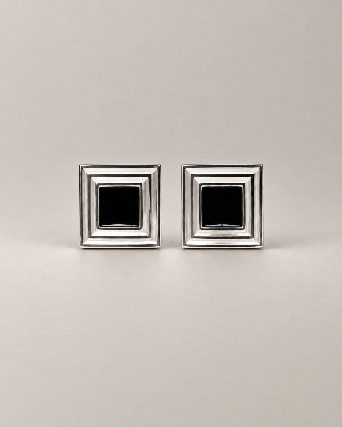 A pair of square silver-toned cufflinks with a black square inset in the center of each, displayed on a neutral light gray background.