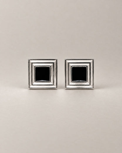 A pair of square silver-toned cufflinks with a black square inset in the center of each, displayed on a neutral light gray background.