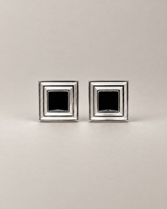 A pair of square silver-toned cufflinks with a black square inset in the center of each, displayed on a neutral light gray background.