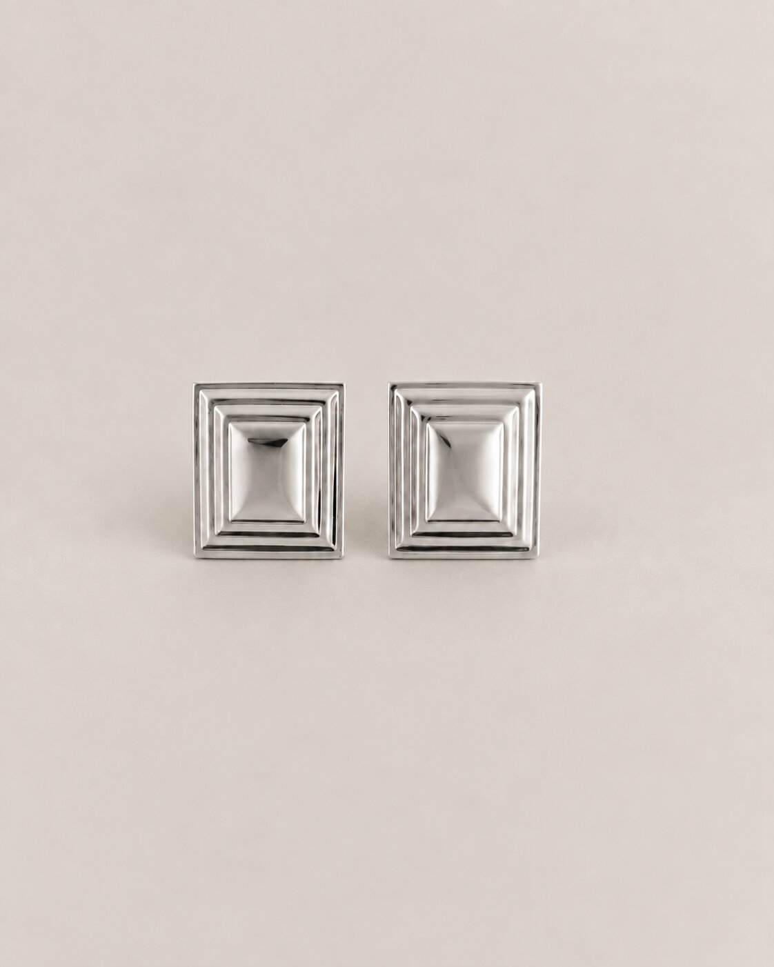 A pair of silver-toned square stud earrings with a stepped, geometric concentric design, displayed on a neutral beige background.