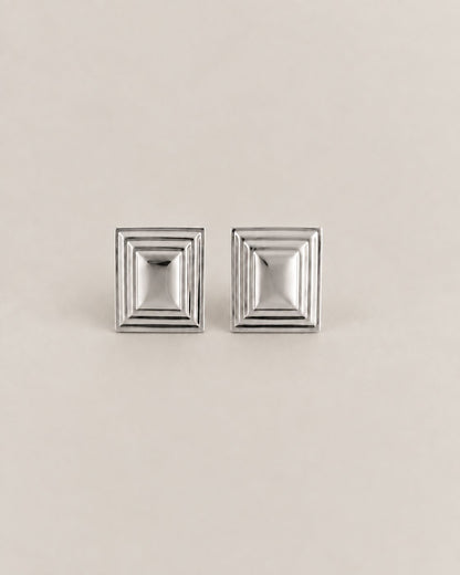 A pair of silver-toned square stud earrings with a stepped, geometric concentric design, displayed on a neutral beige background.