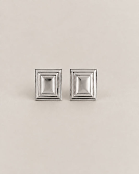 A pair of silver-toned square stud earrings with a stepped, geometric concentric design, displayed on a neutral beige background.