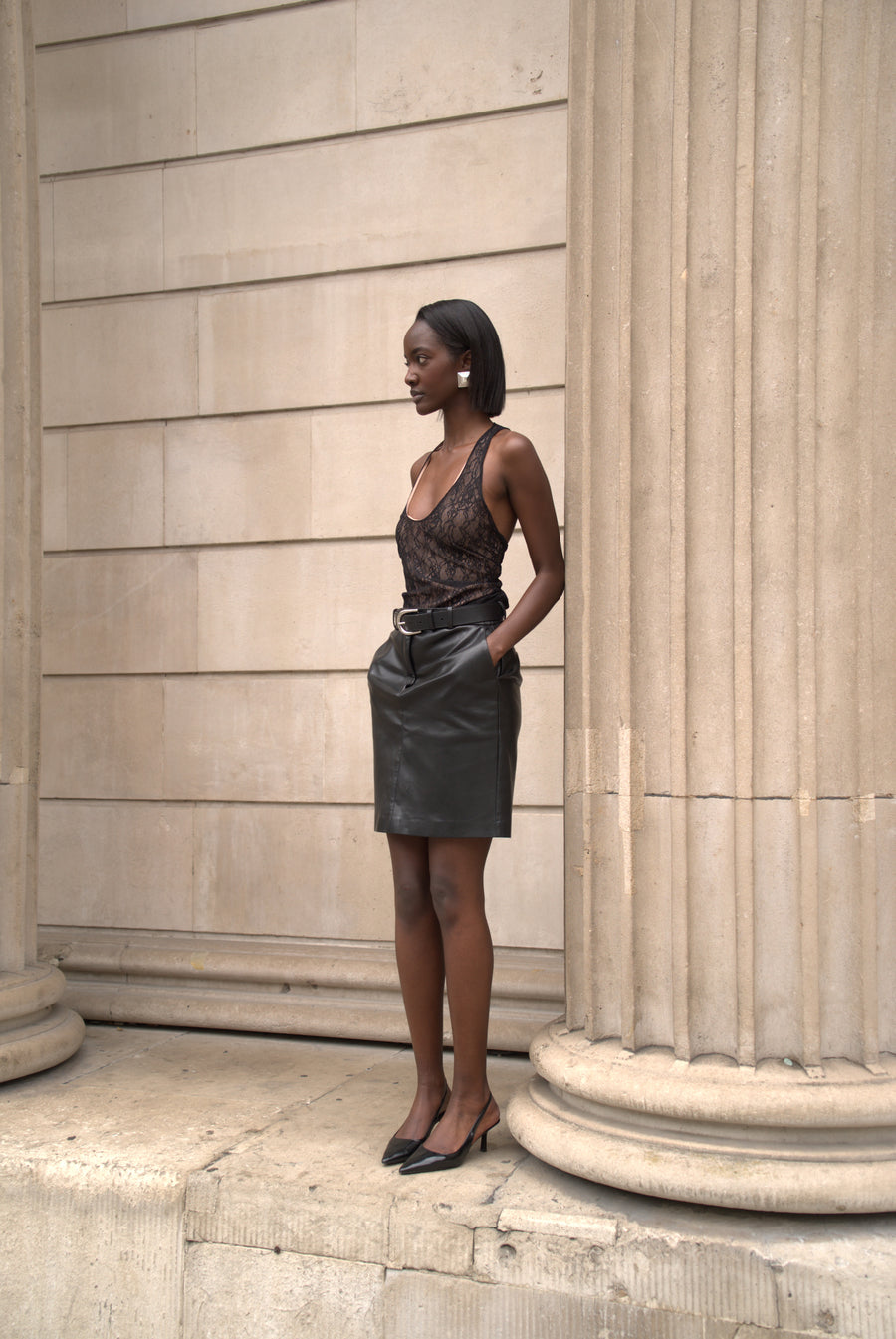 A woman stands outdoors against a beige stone building with large columns, wearing a sheer black lace top and a dark gray mini skirt with a black belt, paired with black pointed flats.