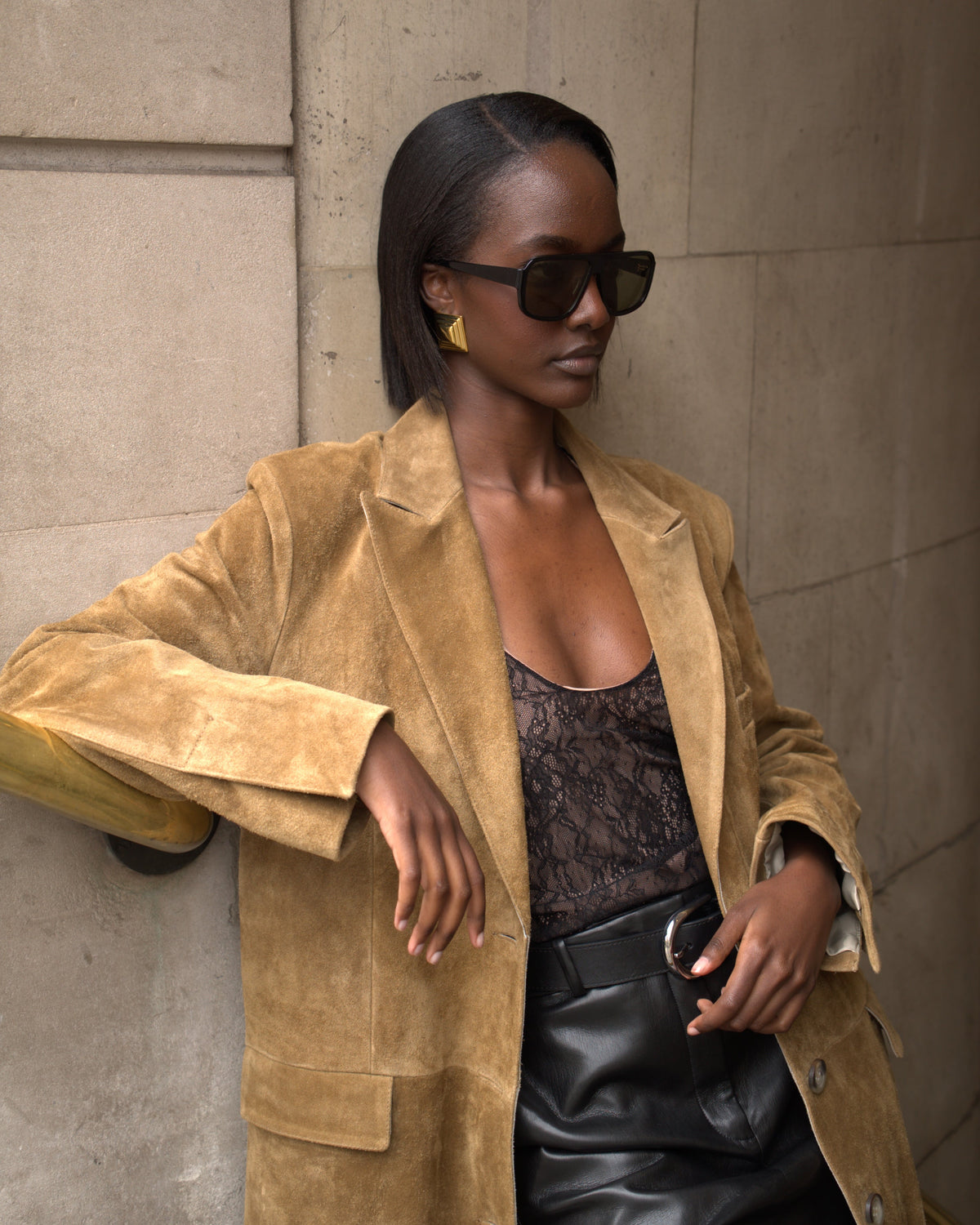 A woman wearing a tan suede blazer over a black lace top and black leather skirt, posing against a stone wall.