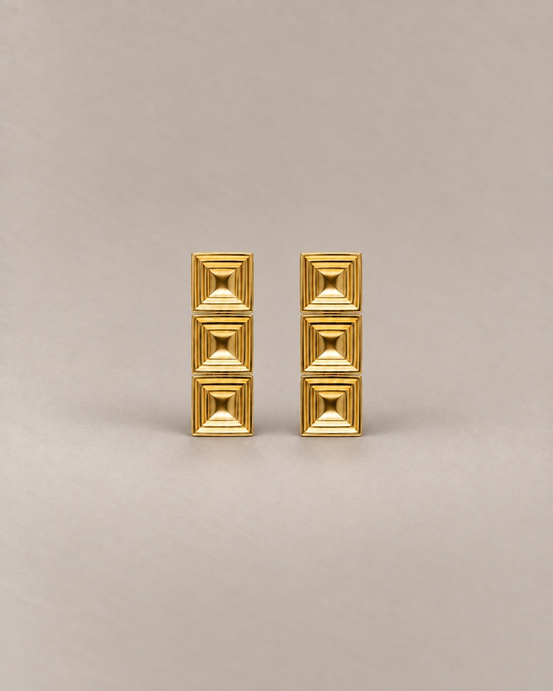 A pair of gold geometric stud earrings with a stacked square pyramid design, set against a solid warm beige background.