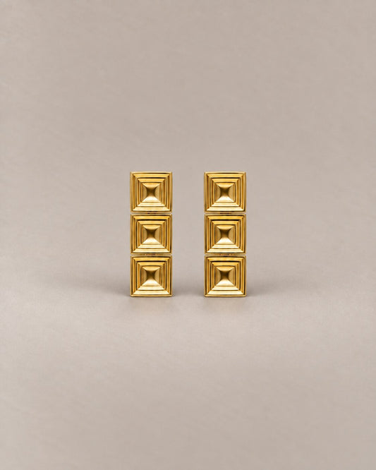 A pair of gold geometric stud earrings with a stacked square pyramid design, set against a solid warm beige background.