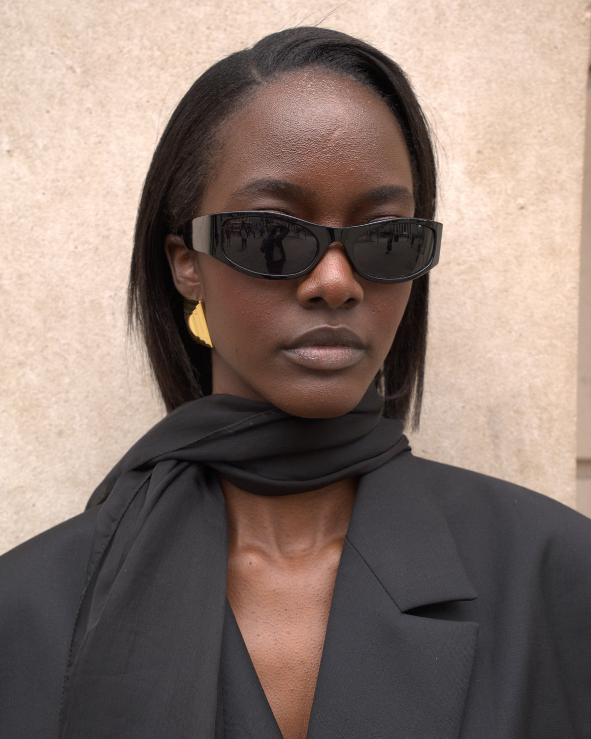 A woman with straight dark hair wears black cat-eye sunglasses, a black scarf, and a black blazer against a beige textured wall.