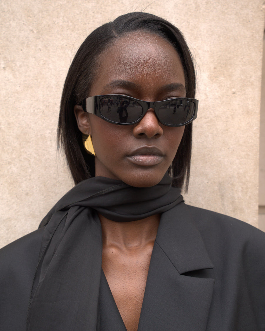 A woman with straight dark hair wears black cat-eye sunglasses, a black scarf, and a black blazer against a beige textured wall.