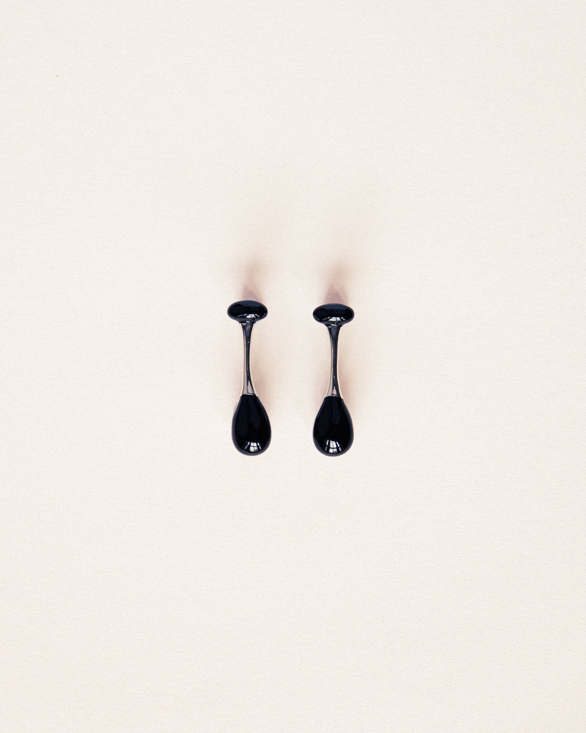 A pair of glossy black teardrop-shaped earrings with thin metallic stems, displayed on a clean, solid off-white background.