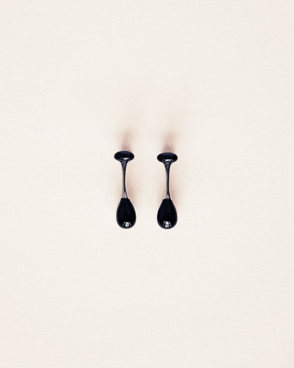 A pair of glossy black teardrop-shaped earrings with thin metallic stems, displayed on a clean, solid off-white background.